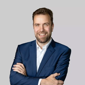 Portrait photo of a man with a full beard, wearing a blue suit and a white shirt. He is looking at the camera with a broad smile, arms crossed, against a neutral grey background.
