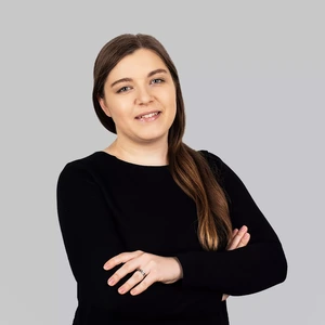 Portrait photo of a woman with long light brown hair, wearing black clothing, smiling at the camera against a light background. Her arms are crossed, and a wedding ring is visible.