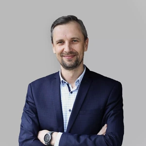 Portrait photo of a man with a full beard, wearing a dark suit and light checkered shirt. He is looking at the camera with a broad smile, arms crossed, against a neutral grey background.